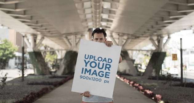 Hipster Dude Holding a Big Poster While Below a Beautiful White Bridge Mockup